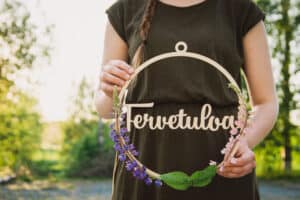 Vihreämekkoinen nainen pitelee lupiineilla koristeltua koivuvanerista ovikranssia jossa teksti tervetuloa.