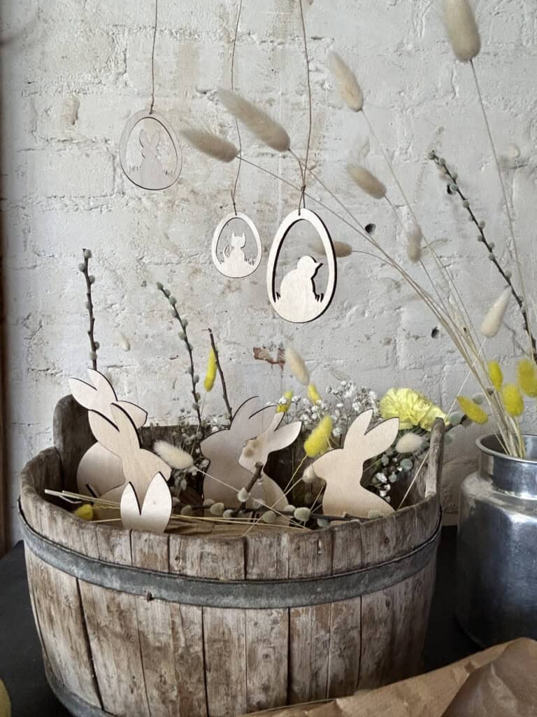 Kuvassa pääsiäisasetelma, jossa on puinen kori täynnä vaneripupuja ja yläpuolella kolme pääsiäismunaa